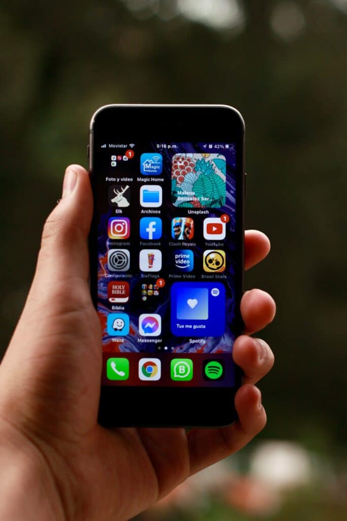 Hand holding iPhone showing app icons: Instagram, Facebook, Clash Royale, YouTube, Spotify, and more.